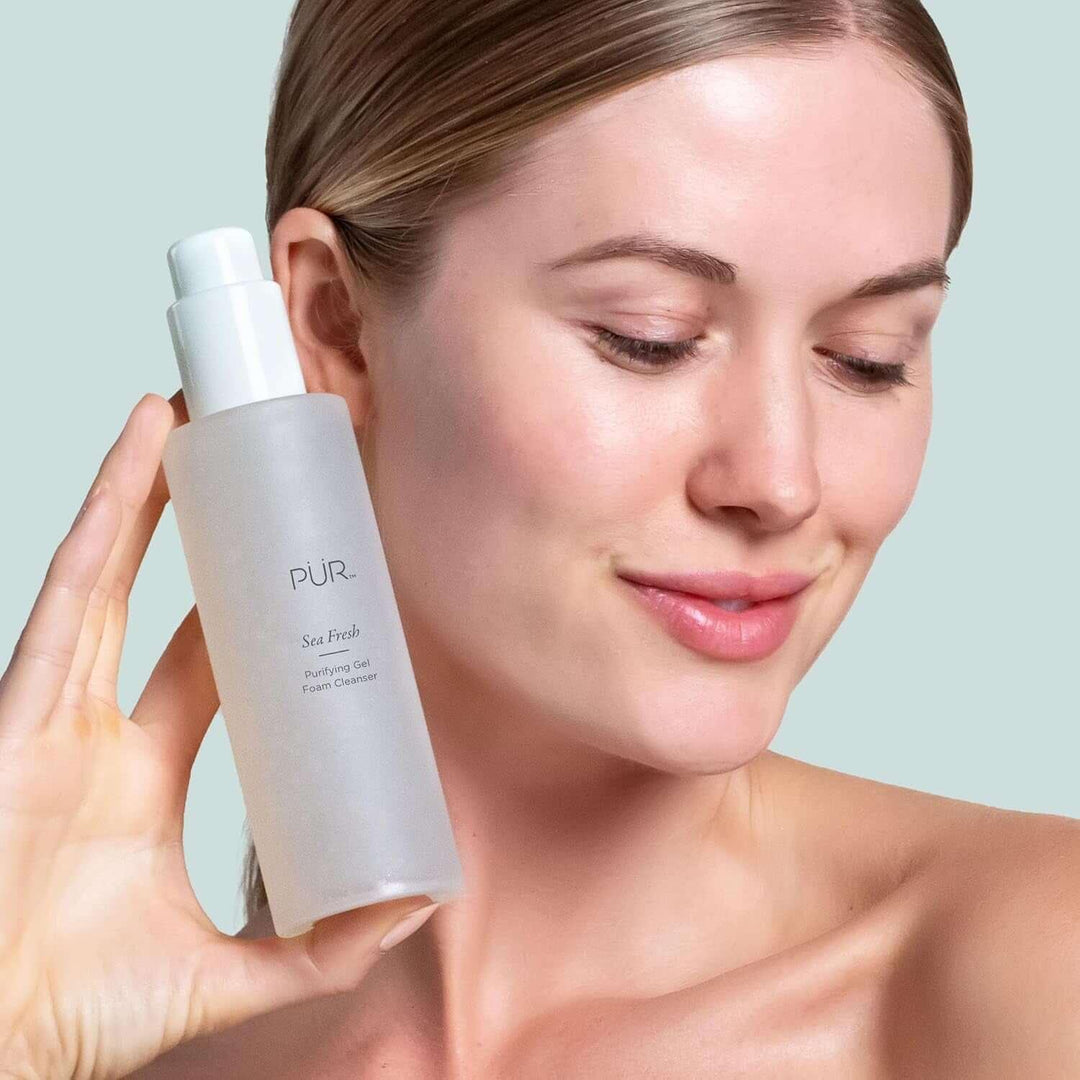 Woman with clear skin holding PÜR Sea Fresh Purifying Gel Foam Cleanser bottle