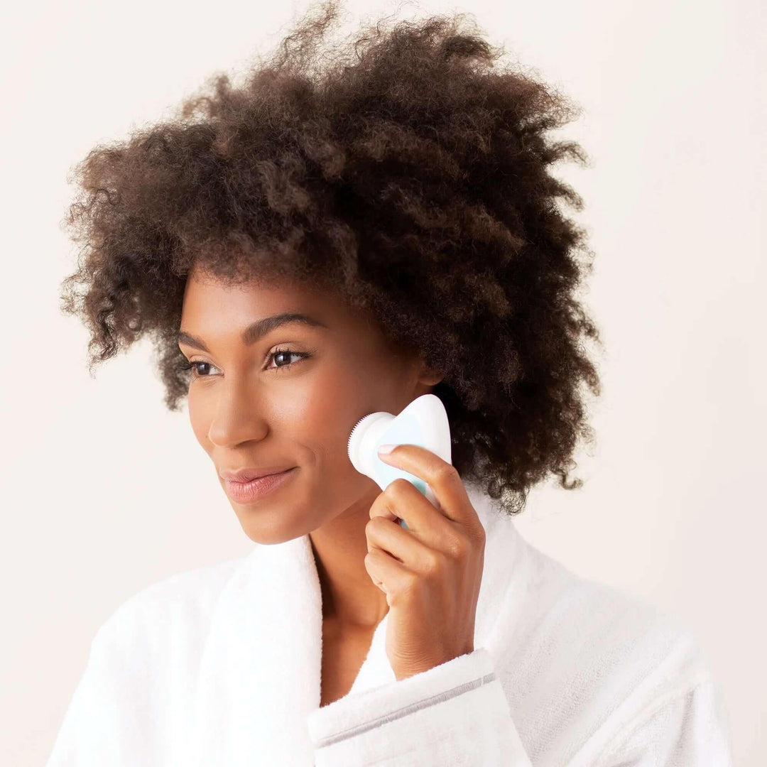Woman using a white facial cleansing brush for skincare, wearing a white robe