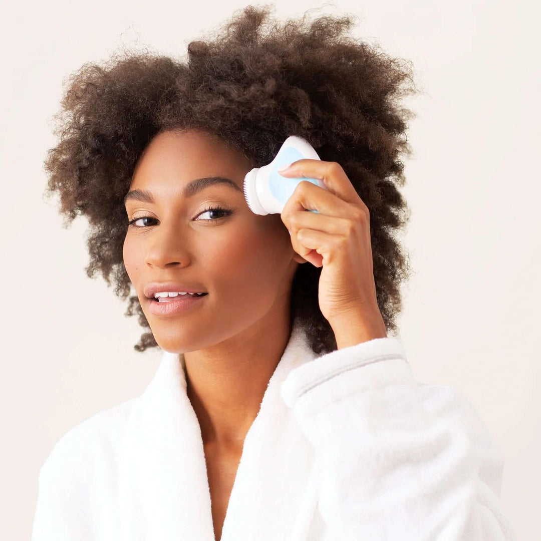 Woman in white robe using electric facial cleansing brush on her cheek