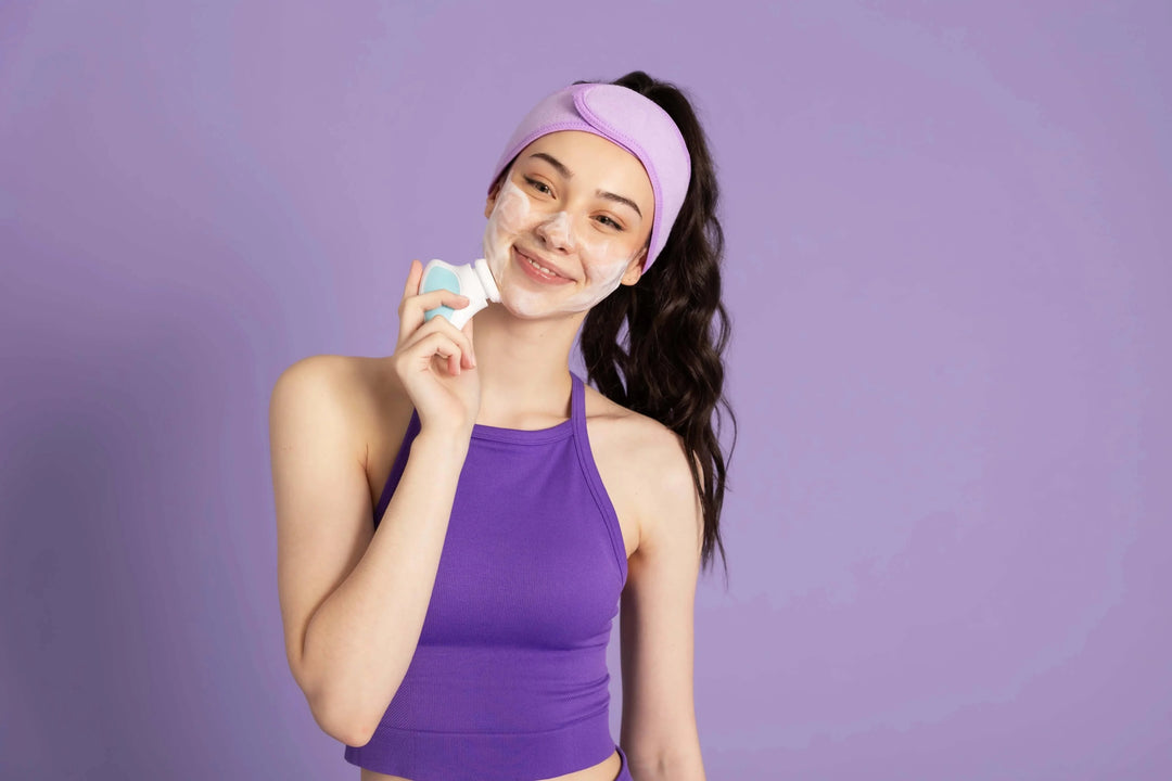 Young woman with foam cleanser on face using facial cleansing brush in purple top and headband