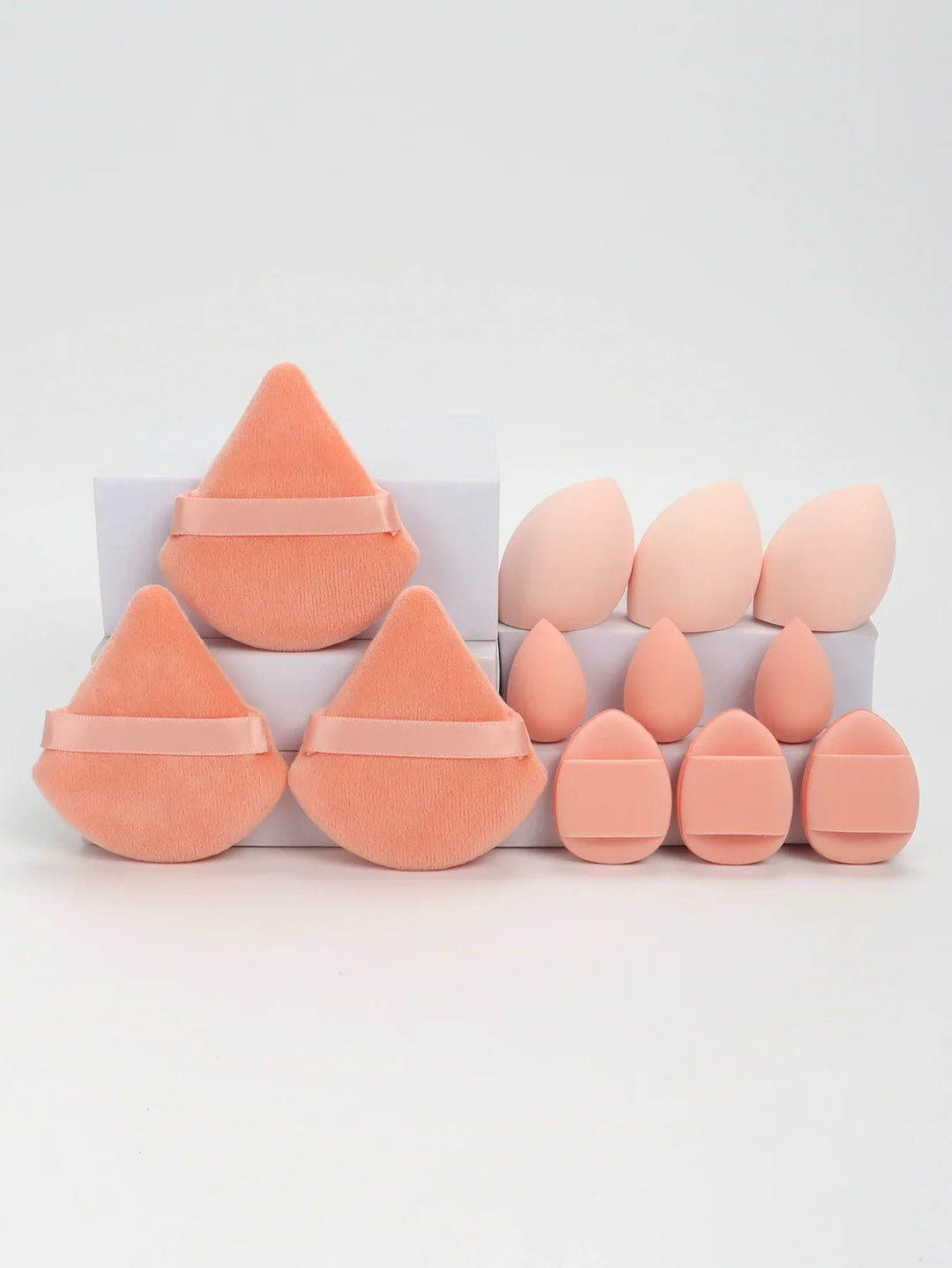 Set of peach-toned makeup sponges and soft powder puffs arranged on white boxes against a white background
