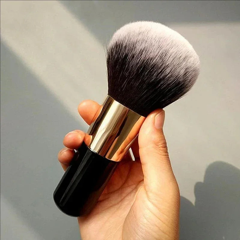 Hand holding a large makeup brush with black handle and dense white-tipped bristles