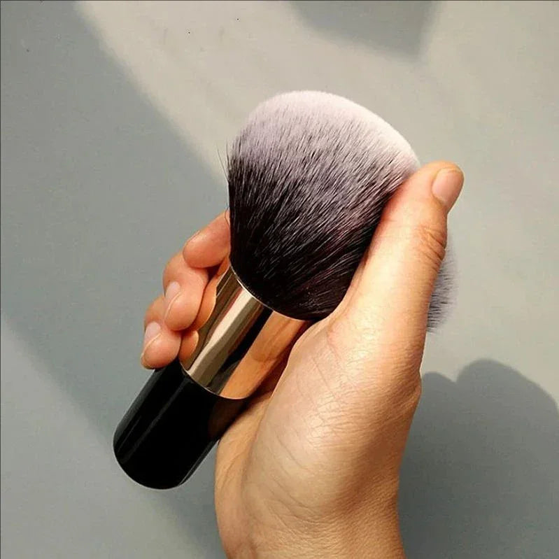 Hand holding a soft, dense makeup brush with black handle and rose gold ferrule against a neutral background