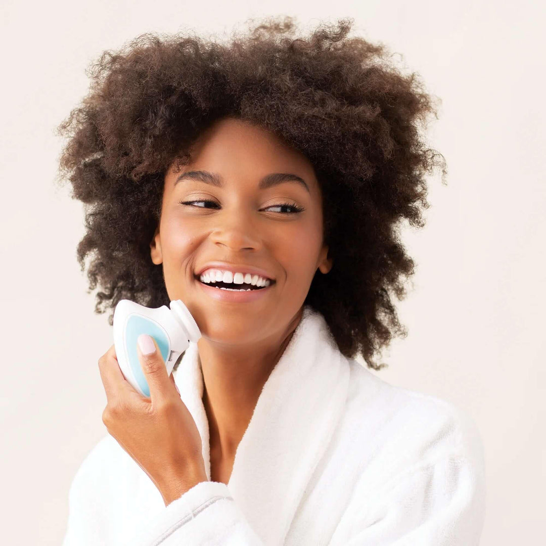 Smiling woman in white robe using electric facial cleansing brush on cheek