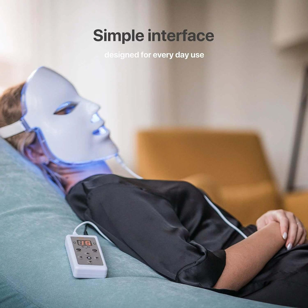 Woman wearing LED light therapy mask with simple interface remote, relaxing on couch