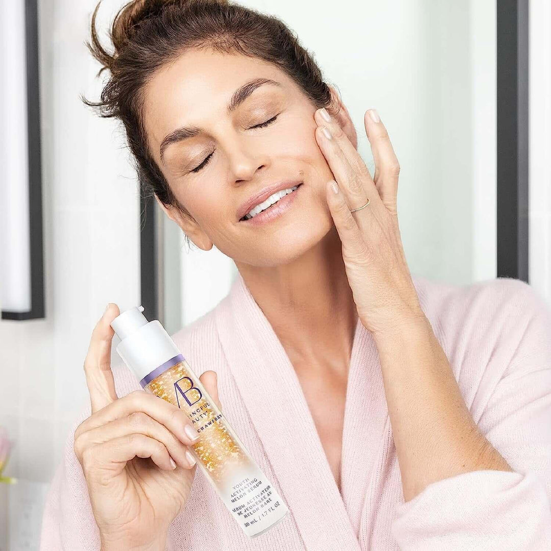 Woman in pink robe applying youth activating melon serum on her face in bright bathroom